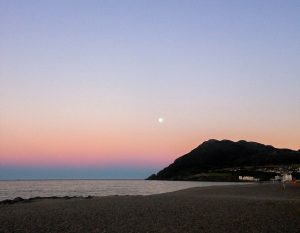 The moon rises over a beach during sunset in Dublin Ireland with mountains in the background.